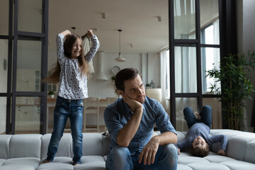 Tired thoughtful father sitting on couch while noisy daughter and son jumping in living room at...