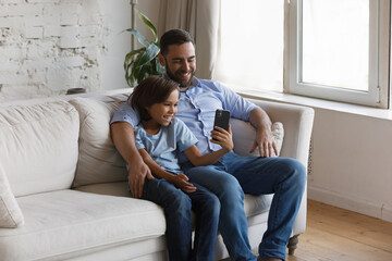 Obraz premium Happy small teen kid boy showing funny video in social network on cellphone to bonding loving young father, posing together for web camera photo or streaming stories online, modern tech addiction.