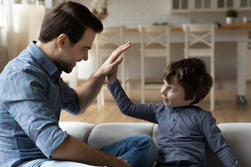 Overjoyed father and little son giving high five, clapping hands, celebrating success, sitting on cozy sofa at home, smiling young Caucasian dad having fun with adorable child boy, playing funny game