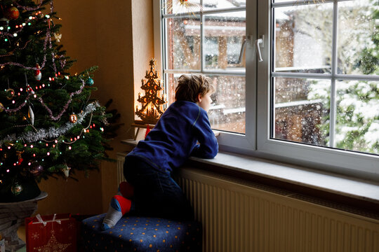 Happy adorable kid boy sitting near window and Christmas tree and looking outside on snow on Christmas day or morning. Smiling healthy child fascinated observing snowfall and big snowflakes