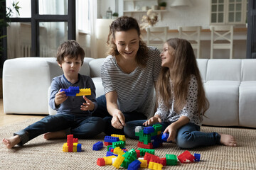 Happy young mother with little son and daughter playing sitting on warm floor in living room at home, smiling mom or nanny and kids building constructing, having fun with colorful plastic blocks