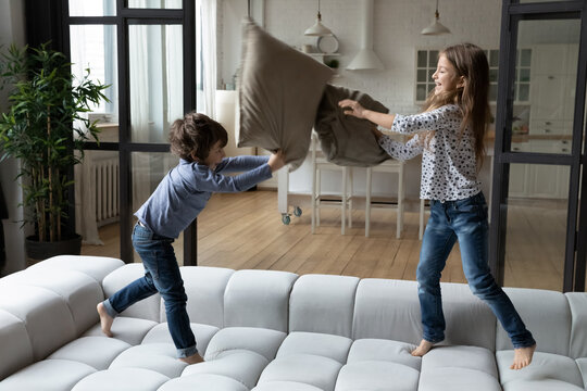 Overjoyed little sister and brother siblings pillow fighting, jumping on cozy couch at home, happy smiling girl and boy having fun, engaged in funny activity, spending leisure time together