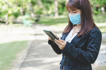 business woman wearing face mask using smartphone tablet in calling for working with her customer in park with sunrise and blur nature background. business new normal with corona virus or covid-19.