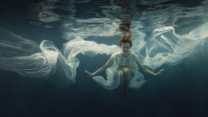 A girl in a white dress posing underwater on a dark background as if she were in zero gravity