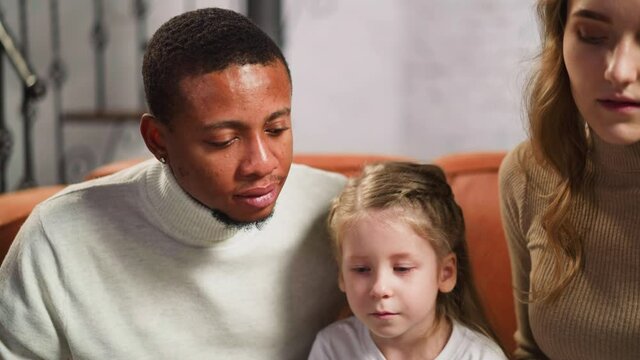 Blonde woman turns page of photobook and little daughter shows grandma picture to mother black colleague in living room closeup slow motion