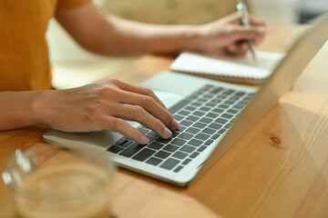 Close-up image of a freelance man taking notes while typing on a computer laptop at the wooden working desk.