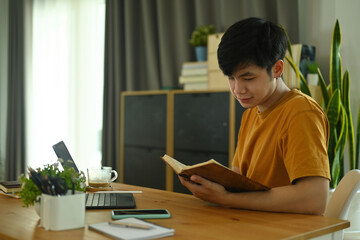 Photo of a young man relaxing with a novel book at the wooden table surrounded by a white blank screen digital tablet and personal equipment.