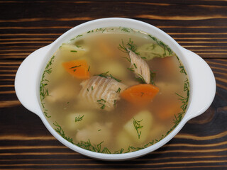 Hot fish soup with vegetables and herbs in a white bowl,  on a wooden table, flat layout. Recipe transparent broth with red fish, carrots and potatoes for a diet lunch