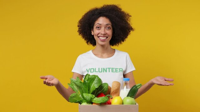 Young black woman 20s wear white t-shirt green title volunteer show on basket with fruit vegetable isolated plain yellow background studio portrait. Voluntary free work assistance help charity concept