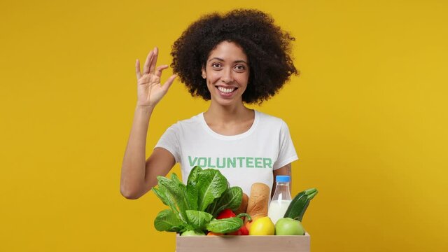 Young black woman 20s in white t-shirt green title volunteer with fruit vegetable basket show okay ok gesture isolated plain yellow background studio portrait. Voluntary free work help charity concept