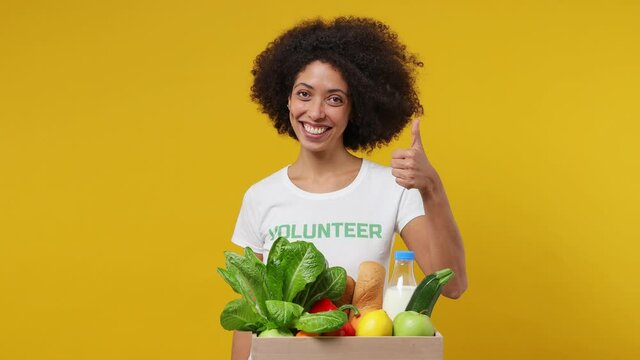Young black woman 20s in white t-shirt green title volunteer point on fruit vegetable basket show thumb up gesture isolated plain yellow background studio portrait. Voluntary free work help concept