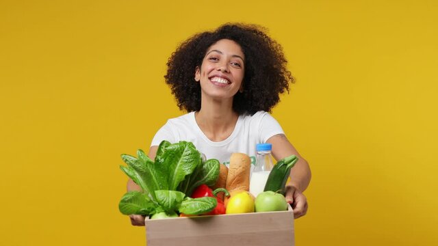 Cheerful young black woman 20s in white t-shirt green title volunteer hold give you fruit vegetable basket isolated on plain yellow background studio portrait. Voluntary free work help charity concept