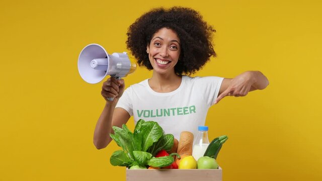 Young black woman 20s in white t-shirt green title volunteer point on fruit vegetable basket scream in megaphone isolated on plain yellow background studio portrait. Voluntary free work help concept