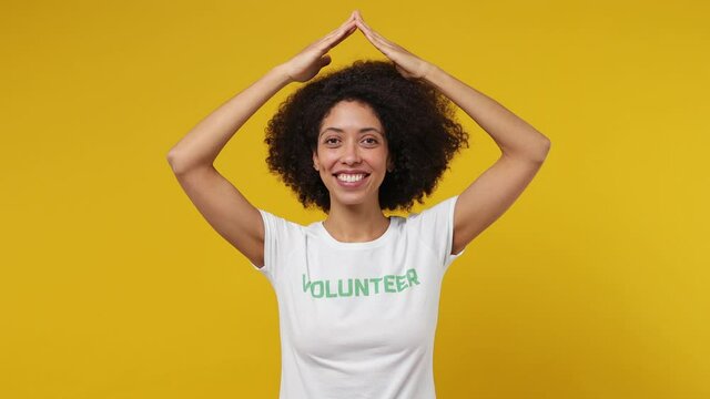 Fun Young Black Woman 20s In White T-shirt Green Title Volunteer Hold Folded Hands Above Head Like House Roof Isolated Plain Yellow Background Studio Portrait. Voluntary Free Work Help Charity Concept