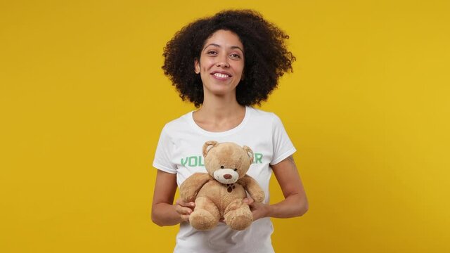 Cute young black woman 20s in white t-shirt green title volunteer hold hug teddy bear plush toy isolated on plain yellow background studio portrait. Voluntary free assistance work help charity concept