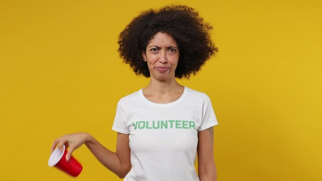 Young black woman 20s in white t-shirt green title volunteer hold red plastic cup say no refuse take paper nod agree yes isolated plain yellow background studio portrait. Stop nature garbage concept