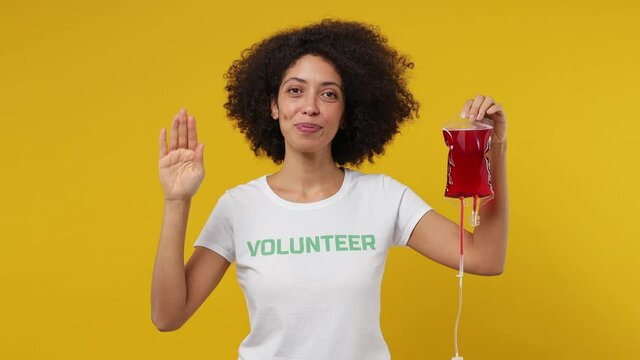 Fun young black woman 20s in white t-shirt green title volunteer hold in hand bag with blood point herself raise hand isolated on plain yellow background studio portrait. Donating transfusion concept