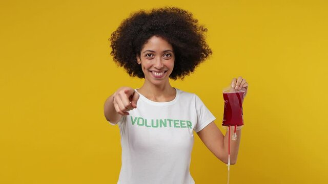 Fun young black woman 20s in white t-shirt green title volunteer hold in hand bag with blood point finger camera you isolated on plain yellow background studio portrait. Donating transfusion concept