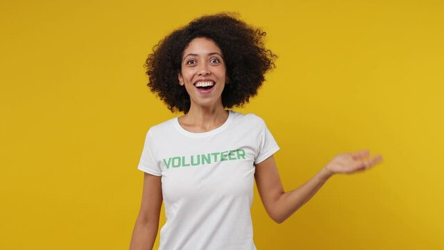 Kind young black woman 20s in white t-shirt green title volunteer appear look search show shape heart sign isolated on plain yellow background studio portrait. Voluntary free work help charity concept