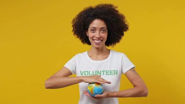 Young black woman 20s in white t-shirt show with palm green title volunteer inscription hold in palms Earth world globe isolated plain yellow background studio portrait. Stop nature garbage concept