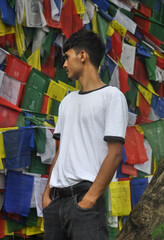 A young man standing outside looking sideways with Buddhist prayer flags in the background © Yaman Kumar 