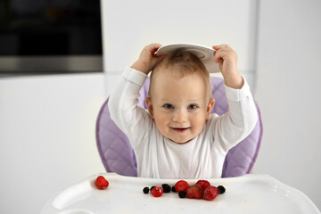 cute blonde baby eats fresh raspberries, strawberries and blueberries in a baby feeding chair. The concept of healthy nutrition for children. High-quality photography