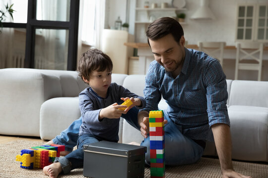 Smiling father and little son playing with colorful plastic construction toys, sitting on warm floor in living room at home, happy loving young dad and child kid spending leisure time together