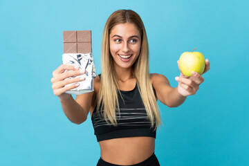 Young blonde woman isolated on blue background taking a chocolate tablet in one hand and an apple...