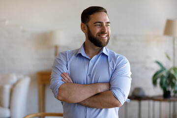 Obraz premium Happy confident young businessman entrepreneur manager standing with folded arms at home office, looking in distance visualizing future, imagining opportunities or thinking of company development.
