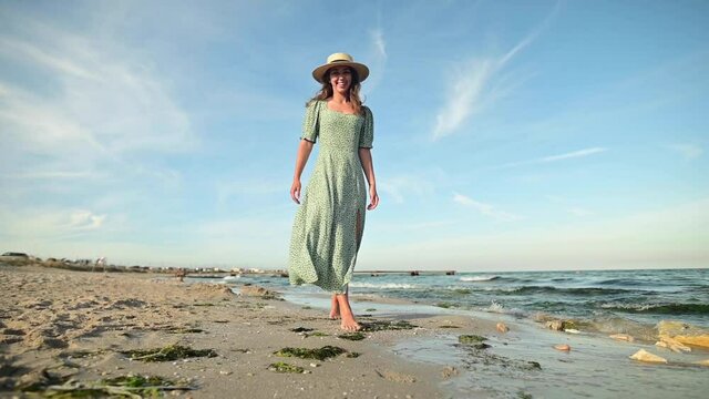 A young attractive Caucasian woman in a straw hat and a green dress is  walking along the coast of the sea on a sunny morning. Romantic girl by the  ocean