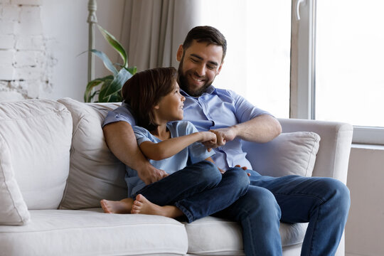 Smiling preteen small kid boy bumping fists with joyful caring young father, making decision, enjoying talking speaking communicating, resting on comfortable sofa together on weekend at home.