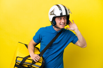 Young Brazilian man with thermal backpack isolated on yellow background listening to something by putting hand on the ear