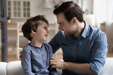 Close up smiling loving dad talking with little son, sitting on cozy sofa at home, happy young...