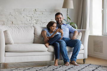 Fototapeta premium Smiling bonding young father and small preteen child son using cellphone, enjoying playing video games, choosing goods shopping in internet store or web surfing, copy space for mobile apps ad.