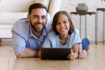 Portrait of happy bonding young father and small preteen kid son using digital computer tablet applications, lying on warm heated floor in living room, spending time online together on weekend.