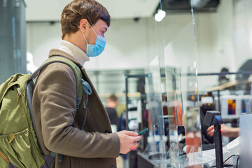 Fototapeta na wymiar Man with short brunet hair in warm sweater coat and medical mask stands near cash register holding phone to pay with contactless payment