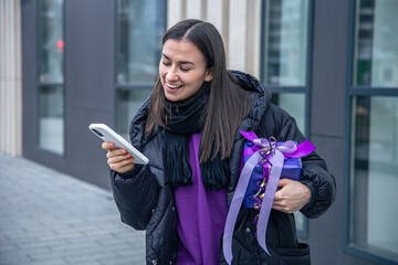 Young woman with a purple gift in hands uses a smartphone outside.