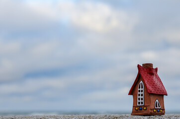 A small ceramic house with a red roof against a gloomy sky on a concrete surface. A dollhouse on...