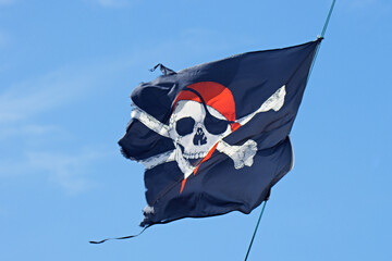 Damaged pirate flag with a skull and cross bones waving in the wind against a blue sky
