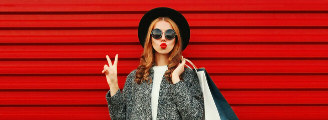 Fototapeta premium Fashionable portrait of stylish young woman model posing with shopping bags and blowing her lips sending sweet air kiss wearing gray coat, hat on colorful red background