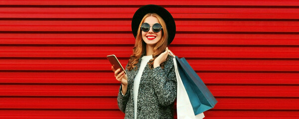 Obraz premium Fashionable portrait of happy smiling young woman with smartphone and shopping bags wearing a gray coat, hat on colorful red background