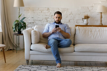 Fototapeta premium Concentrated young man using cellphone software applications, reading news, typing message sending email, web surfing information, playing video game, sitting on couch at home, tech addiction concept.
