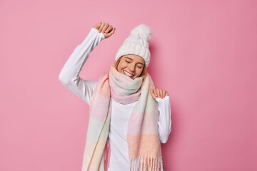 Carefree optimistic woman wears white hat warm winter scarf around neck dances with joyful expression moves with rhythm of music isolated over pink background. Sincere emotions and feelings concept