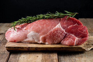 A large piece of raw pork with rosemary lies on a wooden table.