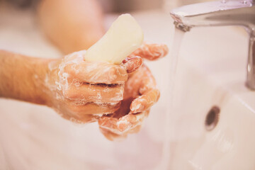 A man carefully washes his hands with soap over a white sink next to the turned on faucet in the bathroom. Cleanliness and hygiene.