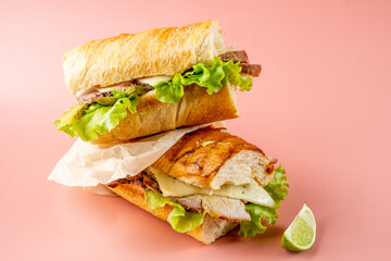 Two cubano sandwiches with baked meat and cheese and pickled cucumbers on a pink background.
