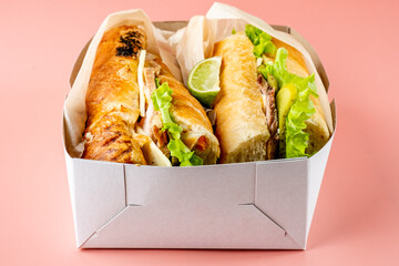 Two sandwiches with meat in a blush baguette in a paper box for delivery. Cubano sandwiches with meat and pickles with mustard sauce.