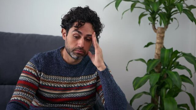 Portrait of handsome man wearing a shirt and feeling exhausted on a couch at home. Frustrated caucasian man holding head on hand and sitting on sofa of house.