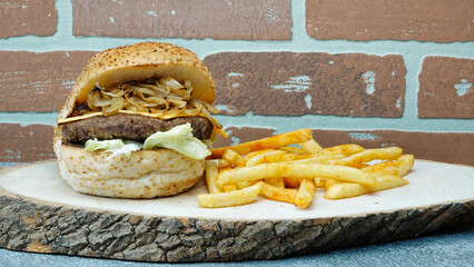 big juicy hamburger with caramelized onions, cheddar cheese, french fries, lettuce on wooden tray with brick wall background	