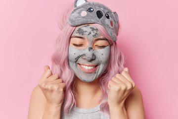 Joyful woman clenches fists with happiness rejoices something keeps eyes closed applies anti aging beauty clay mask wears sleepmask poses against pink background has long dyed hair. Skin care
